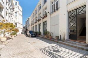 A narrow cobblestone street lined with multi-story buildings, a parked car, and potted plants.