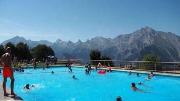 Ein Schwimmbecken mit Schwimmern und spielenden Personen, umgeben von Bergen.
