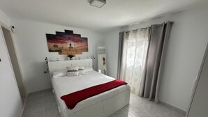 Quarto com cama branca, manta vermelha, pintura na parede e janela com cortinas.