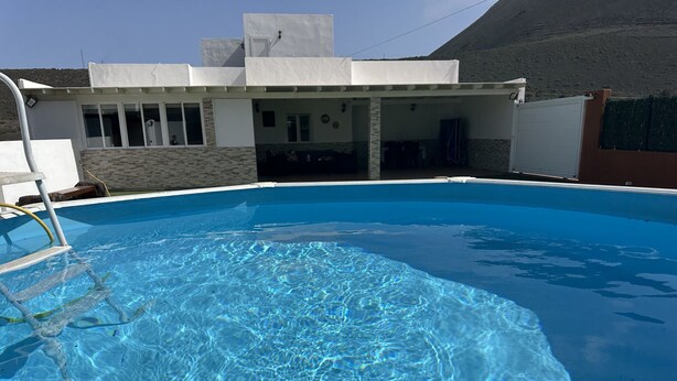 Uma piscina com superfície de água azul cristalina, uma escada e um edifício com área de varanda coberta ao fundo.