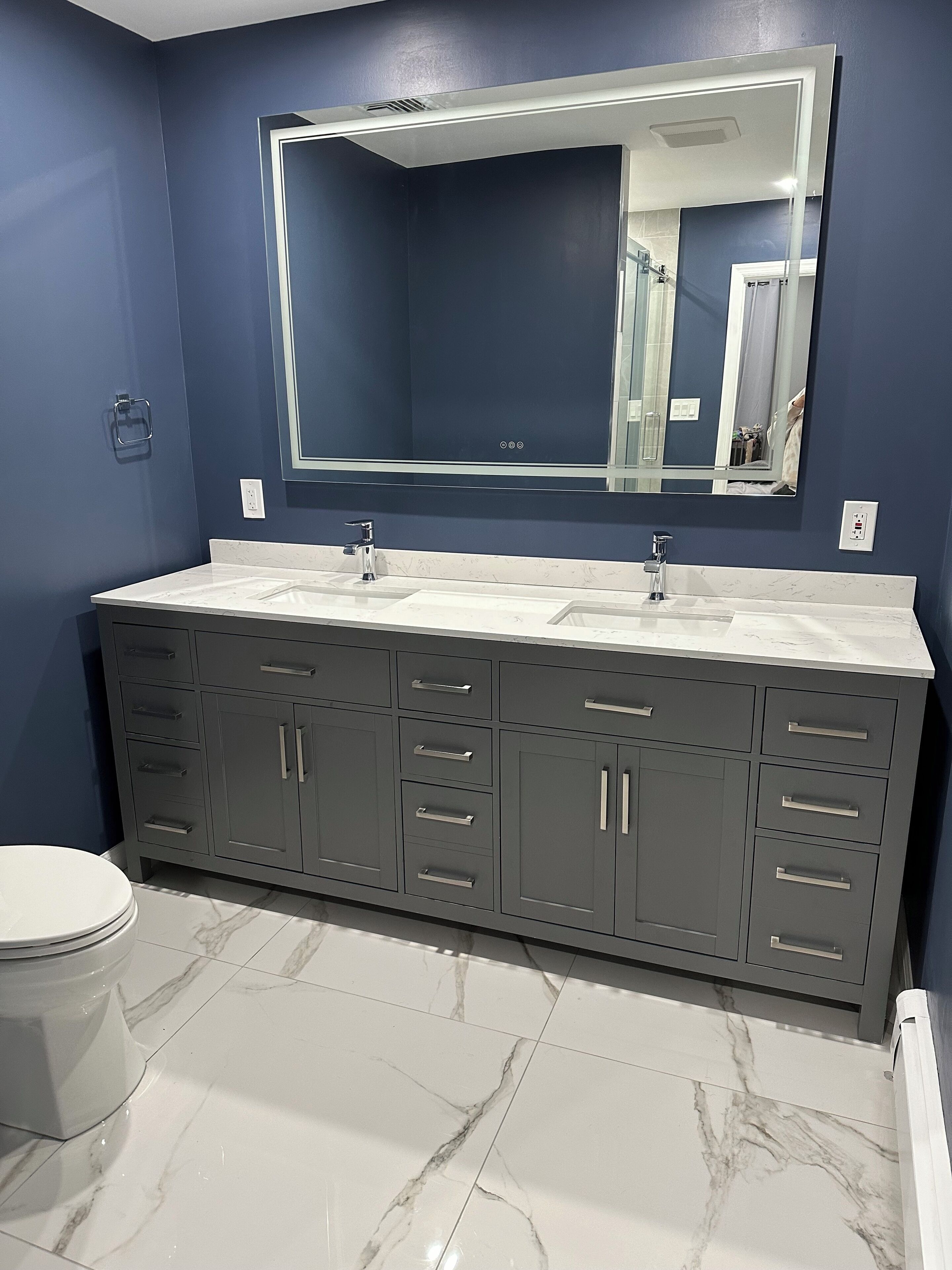 A bathroom with a dark blue wall, a large mirror, a white countertop, and a grey cabinet with multiple drawers.