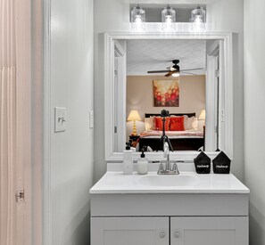 Une salle de bain avec un meuble-lavabo blanc, un miroir, un ventilateur de plafond et un lit visible dans le reflet.