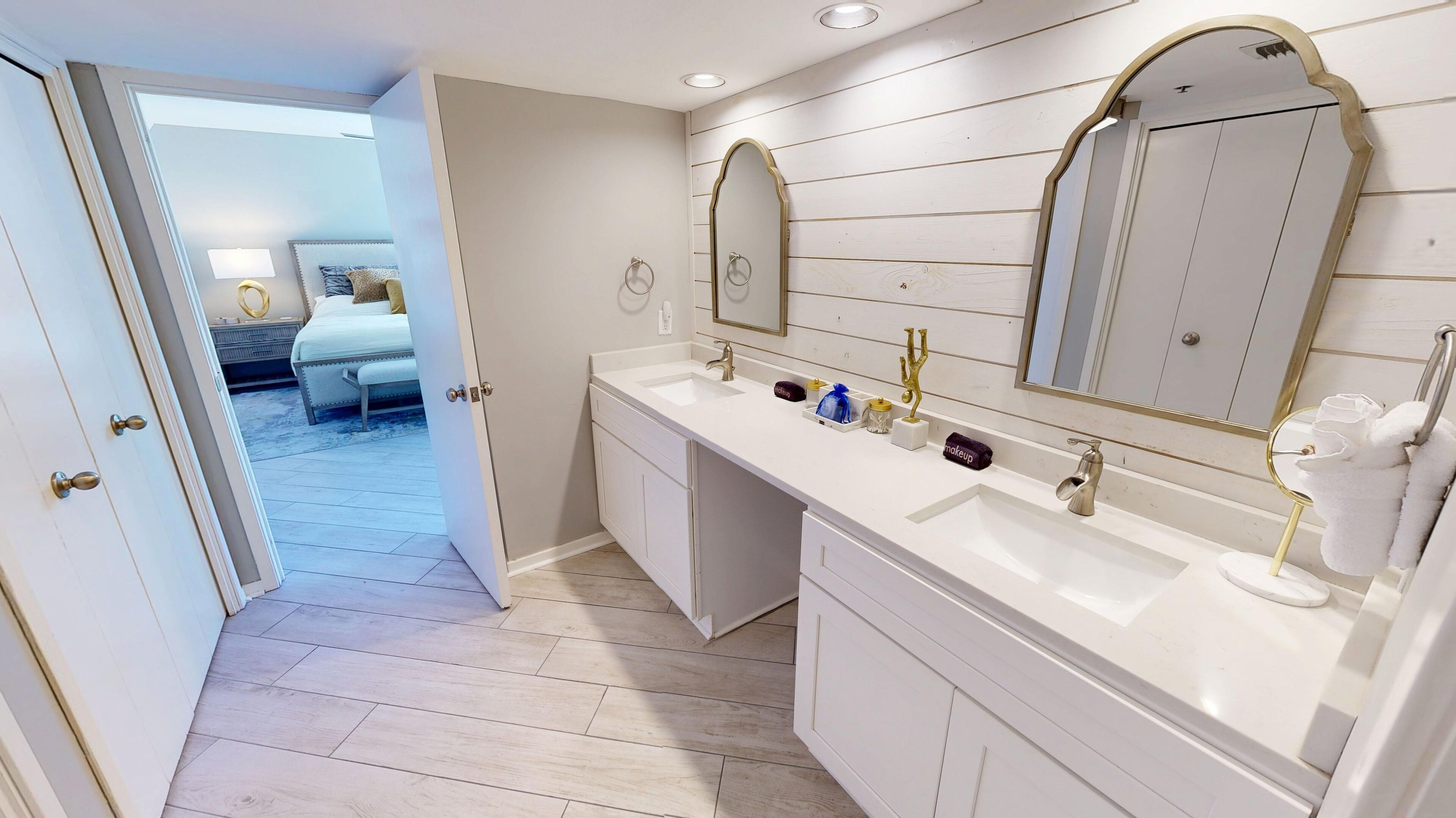 Ein modernes Badezimmer mit einem weißen Waschtisch, zwei Waschbecken und großen Spiegeln. Ein Bett ist im angrenzenden Zimmer zu sehen.