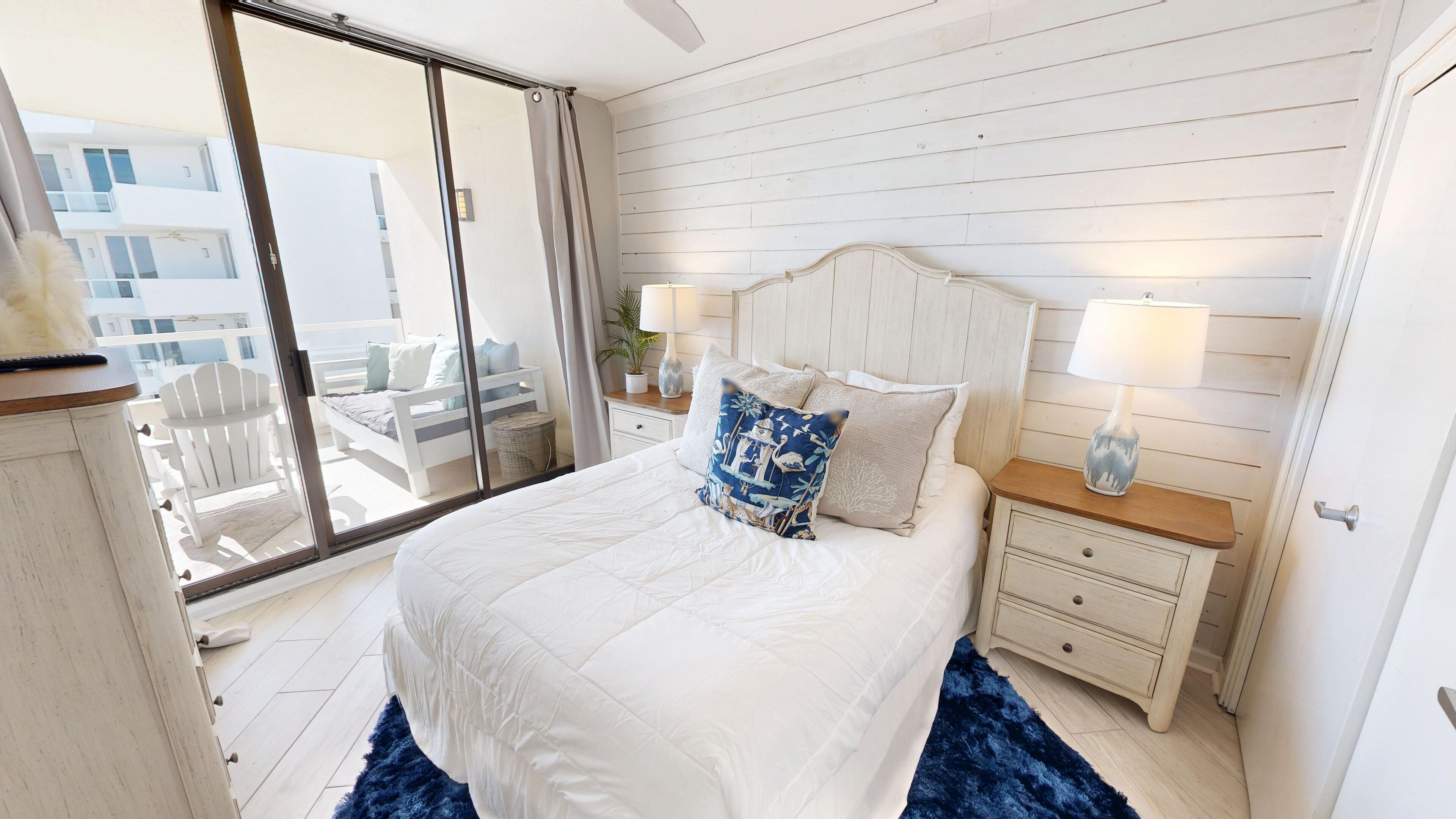 A bedroom with a large bed, a nightstand with lamps, a view through a sliding door, and a balcony with outdoor furniture.