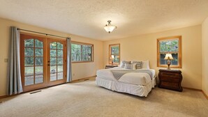 A bedroom with a large bed, a nightstand with a lamp, and a window with a view of trees. - 4 Tan Oak Lane 5 Bedroom Home (Bend)