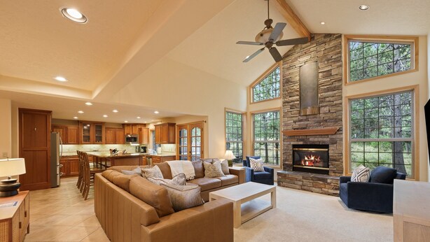 A spacious living area with a stone fireplace, large windows, and a kitchen in the background. - 4 Tan Oak Lane 5 Bedroom Home (Bend)
