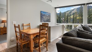 Uma área de refeições com mesa e cadeiras de madeira, um quadro na parede e vista para montanhas através de grandes janelas.