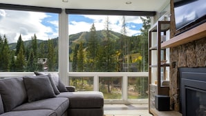 Uma sala de estar moderna com um sofá seccional cinza, uma lareira de pedra e uma grande janela com vista para as montanhas.