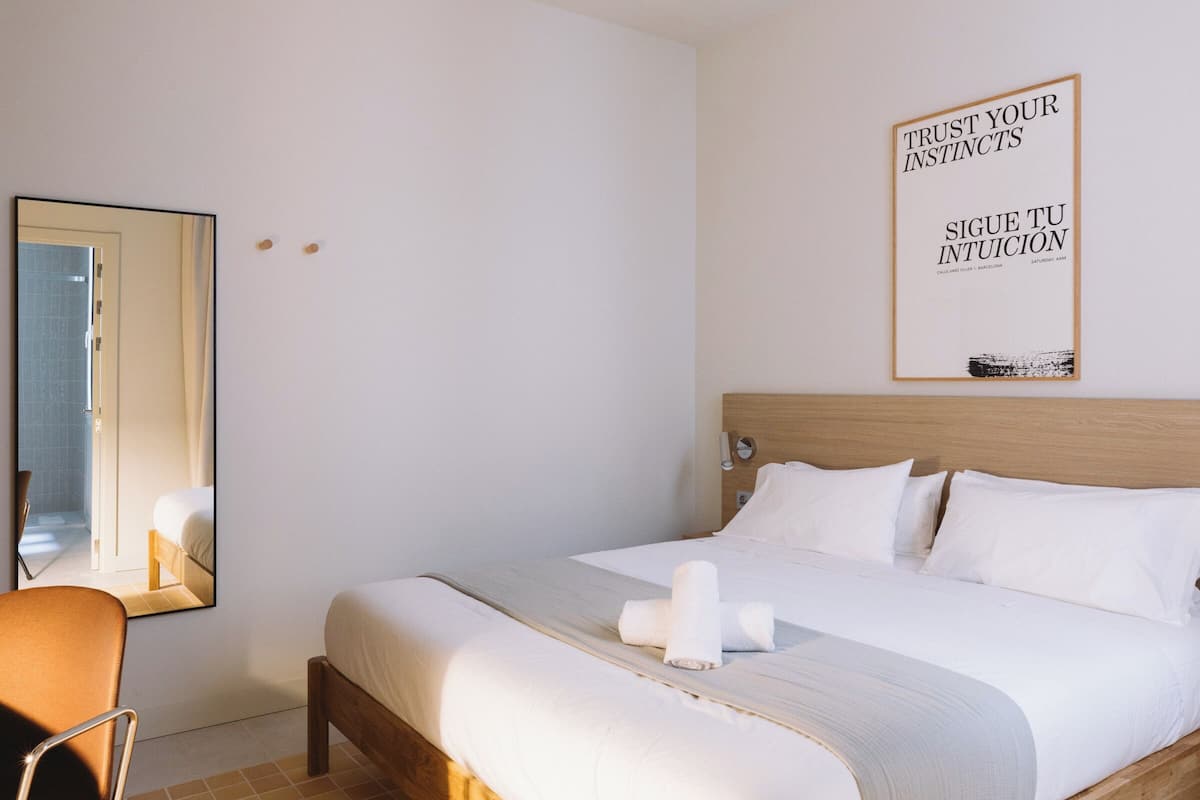 A neatly made bed with white linens and a wooden headboard, a bedside table with a lamp, and a wall-mounted mirror.