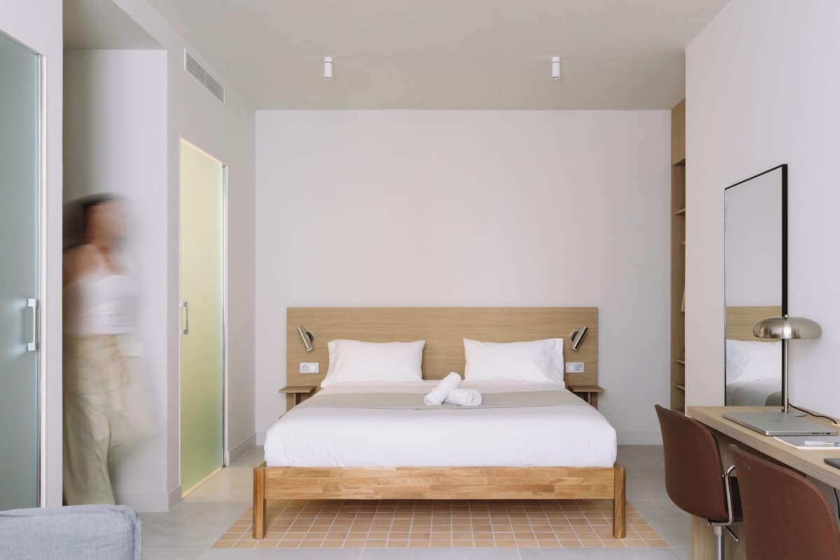 A modern hotel room with a wooden bed, a desk with a chair, a mirror, and a door leading to another room.