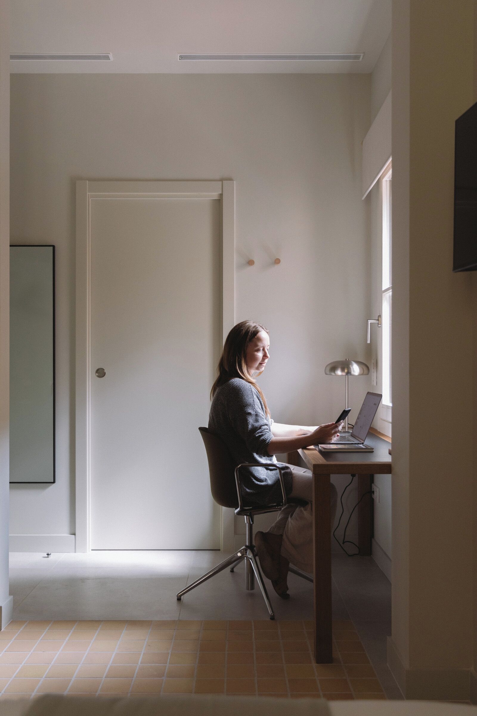 A person seated at a desk with a laptop and smartphone, in a room with a door and a window.