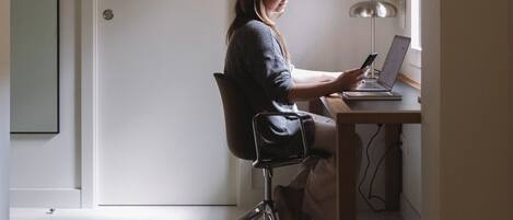 A person seated at a desk with a laptop and smartphone, in a room with a door and a window.