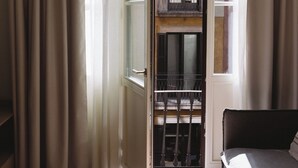 A room with a bed, a round table with a remote control, a window with curtains, and a view of a building with balconies. - Outsite Barcelona - Work, Connect, Explore (Barcelona)