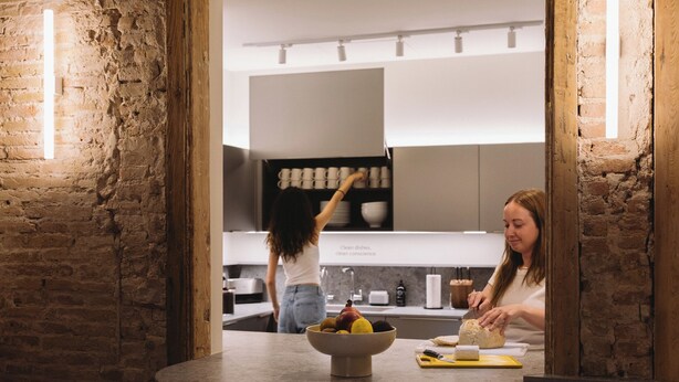 A kitchen with a rustic brick wall, a bowl of fruit on the counter, and a person preparing food. - Outsite Barcelona - Work, Connect, Explore (Barcelona)