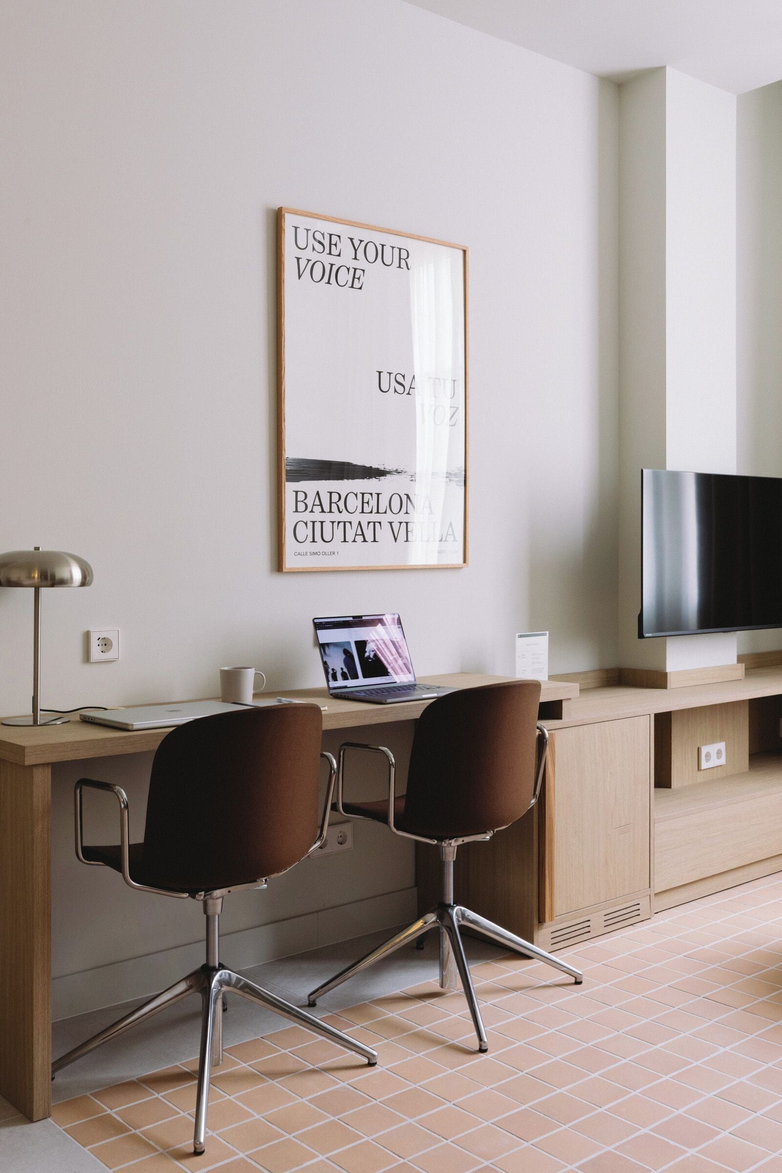 Une chambre d’hôtel moderne avec un bureau en bois, deux chaises, un ordinateur portable, un téléviseur à écran plat et une affiche motivante accrochée au mur.