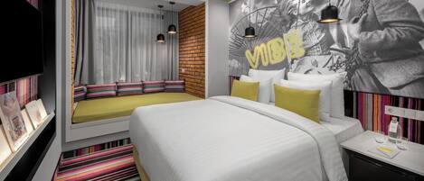 A modern hotel room with a large bed, a seating area with cushions, a TV, and a wall mural.