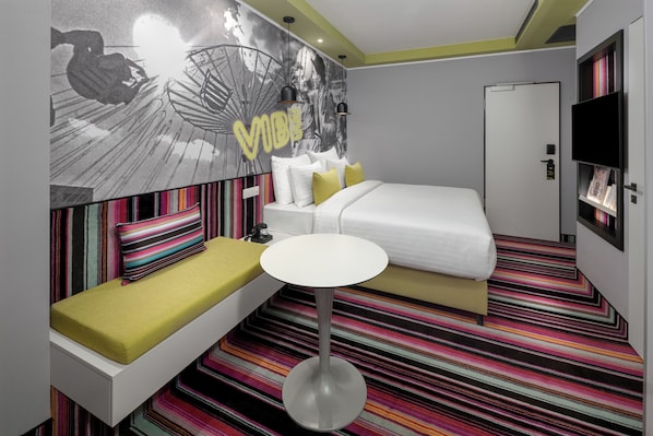 Habitación de hotel moderna con una cama, una mesita pequeña redonda, un banco con cojines, una alfombra colorida a rayas y un mural en la pared.