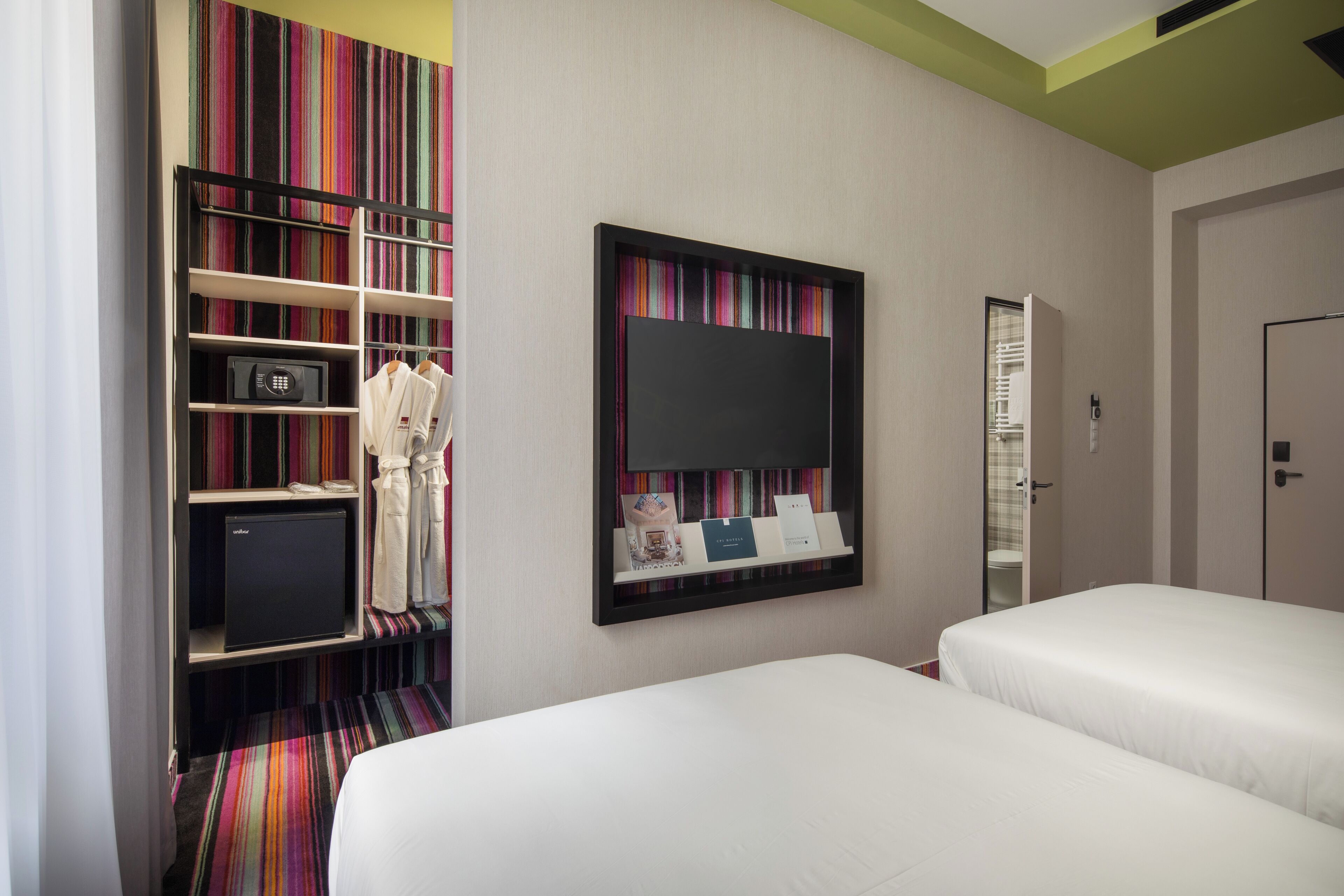 Habitación de hotel con dos camas, un armario con batas, un refrigerador pequeño y una estantería con libros y un espejo.