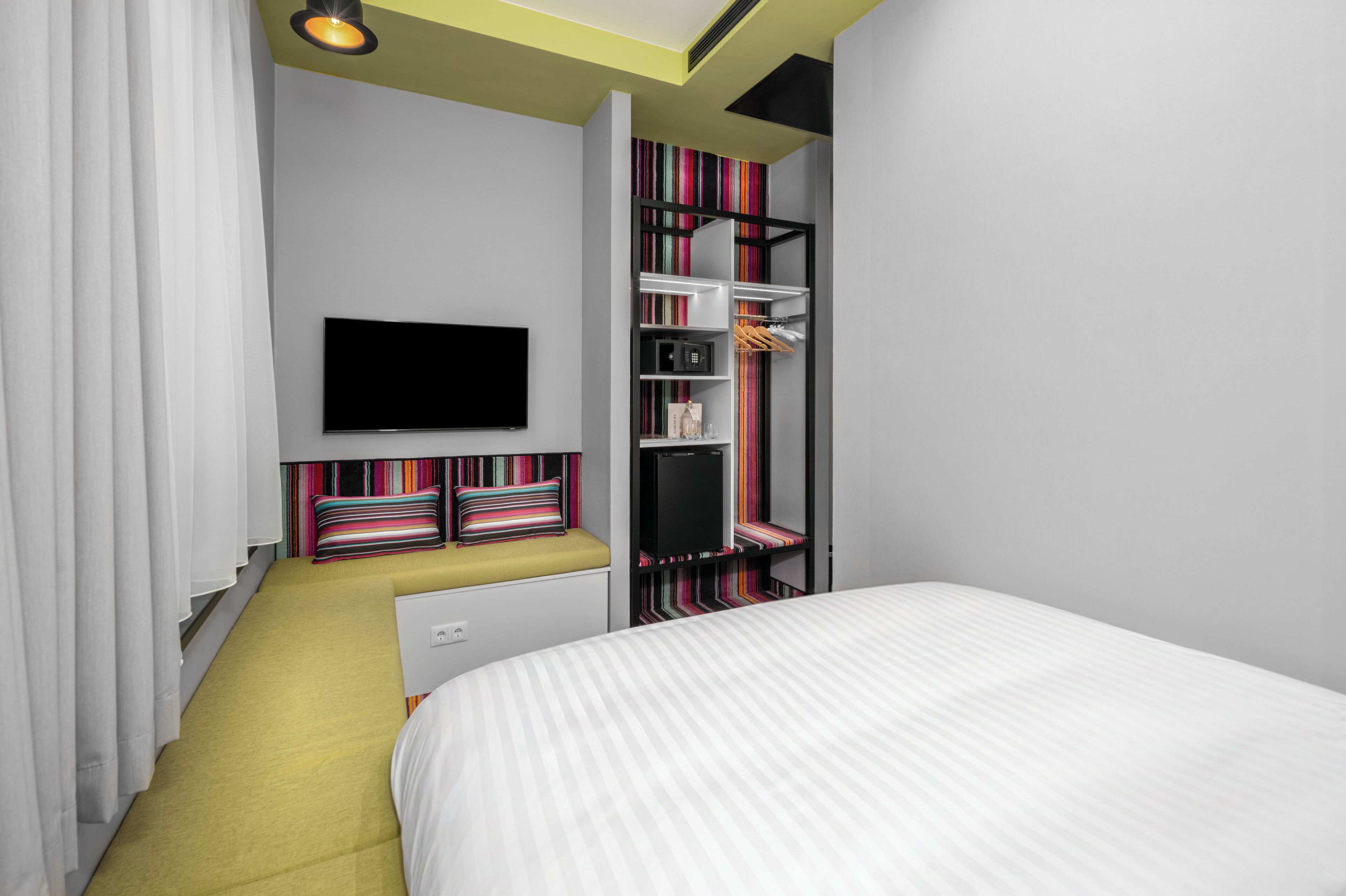 Una habitación de hotel moderna con una cama, un televisor montado en la pared, un armario empotrado y un banco con cojines de colores.