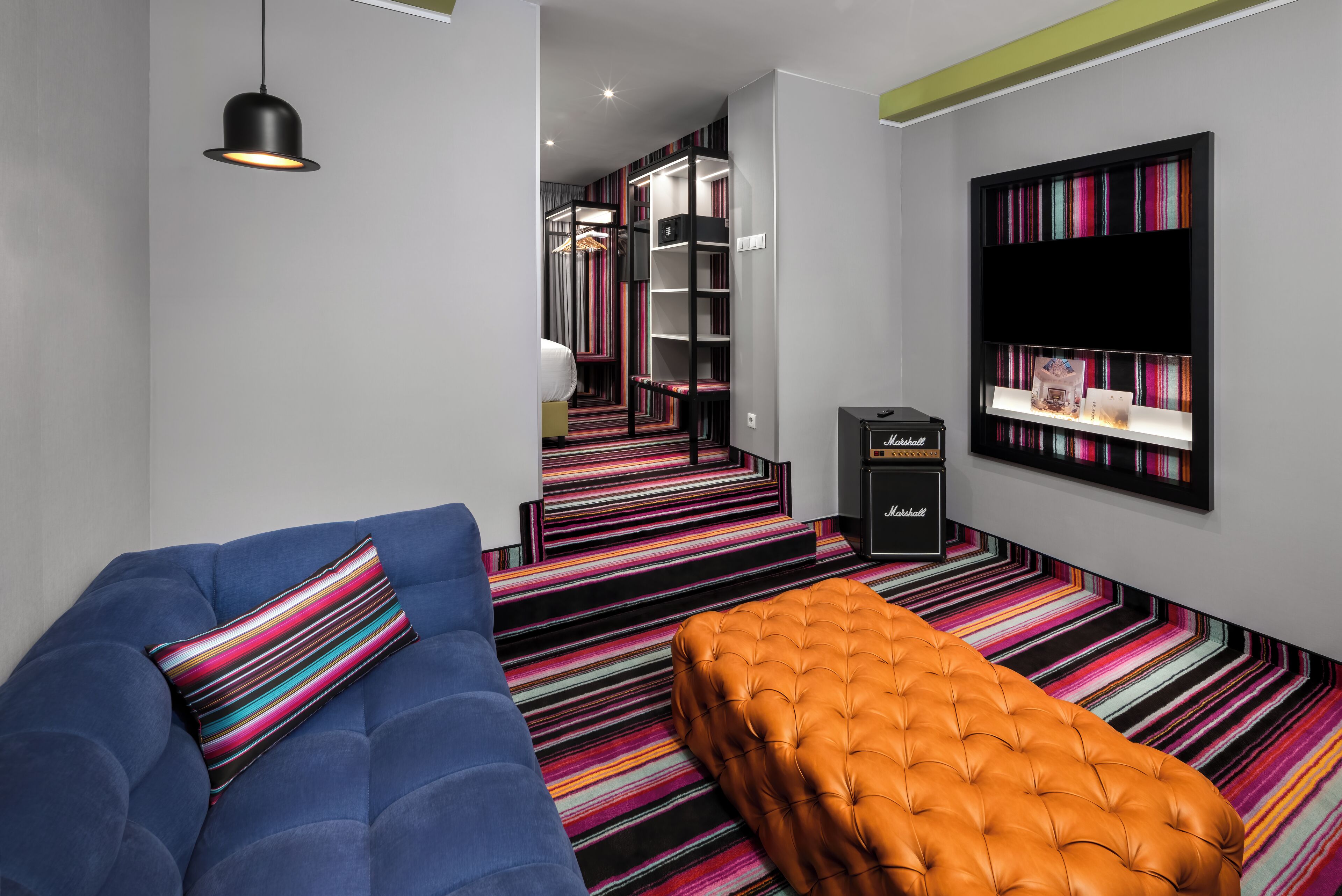 Una habitación de hotel moderna con un sofá azul, un otoman naranja, una alfombra a rayas y una estantería con libros.