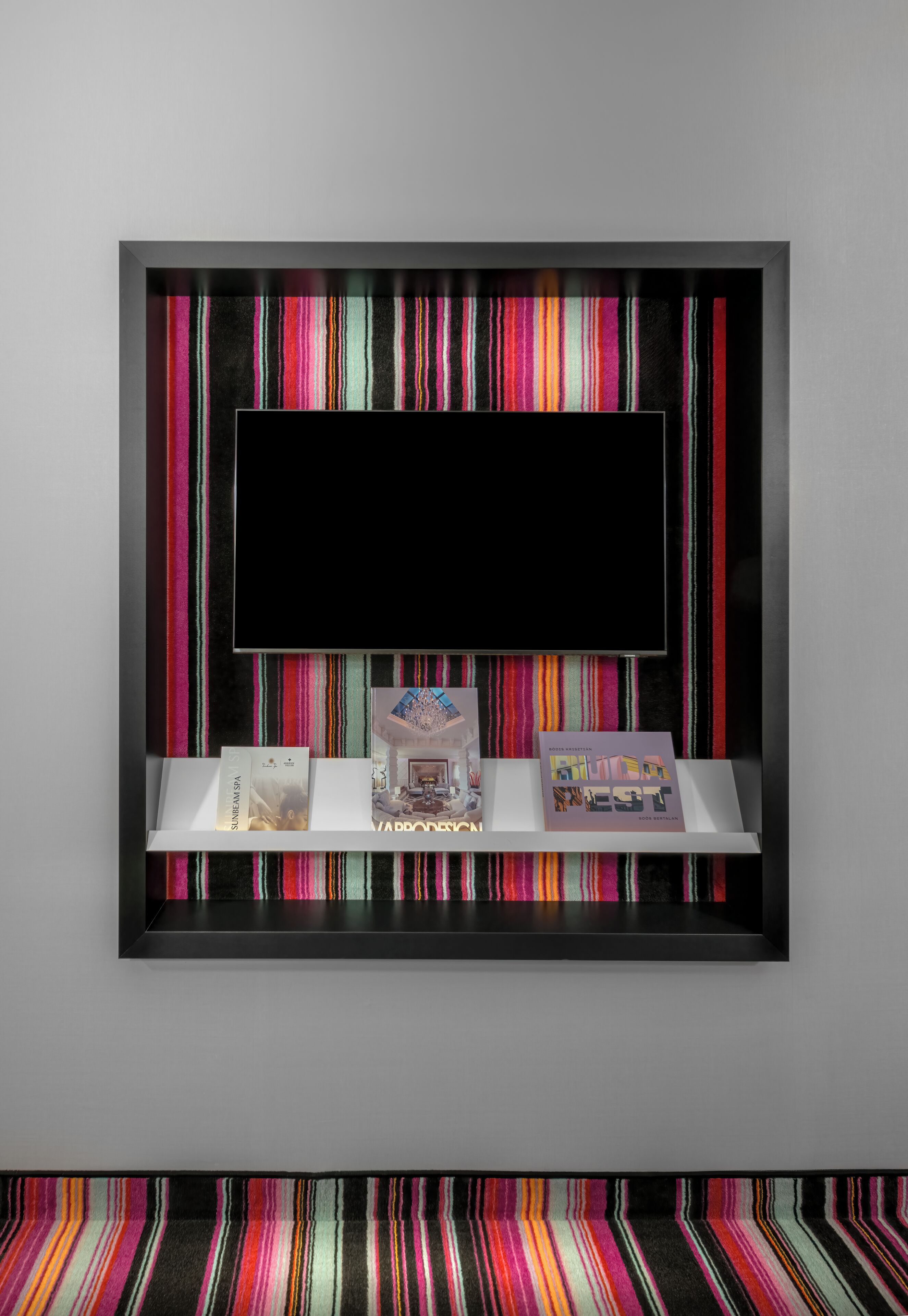 Una camera moderna con un'ampia televisione montata su una parete rivestita con carta da parati colorata a righe. Sotto la TV, si vedono tre libri su uno scaffale.