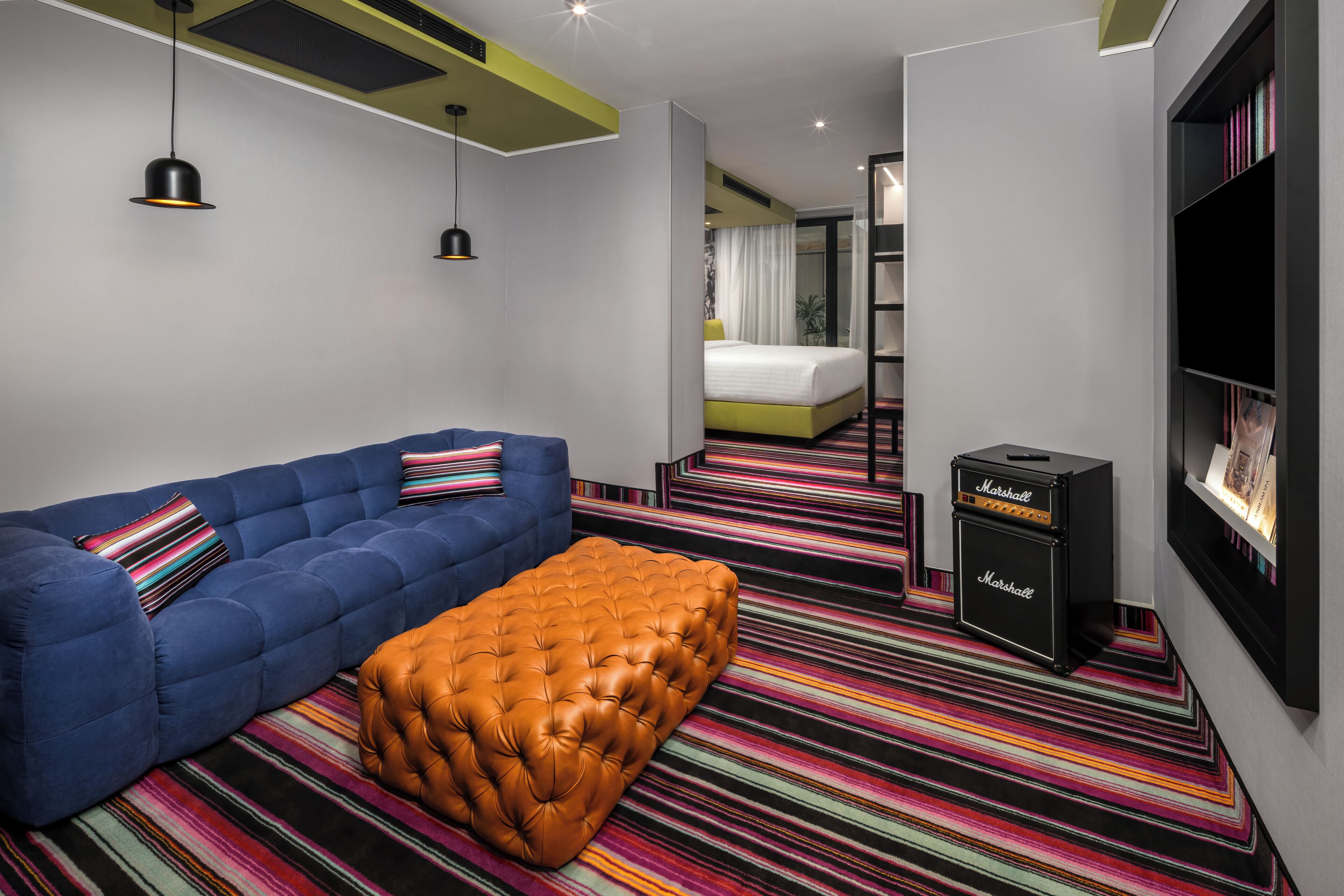A modern hotel room with a blue sofa, a brown ottoman, a striped carpet, a bookshelf, and a Marshall amplifier.