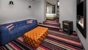 A modern hotel room with a blue sofa, a brown ottoman, a striped carpet, a bookshelf, and a Marshall amplifier.