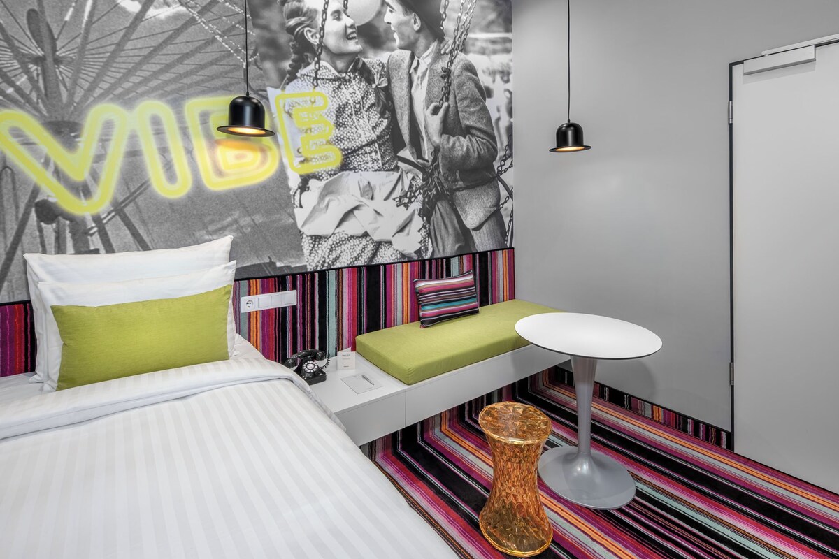 Una camera d'albergo moderna con un letto, un comodino con una lampada, un piccolo tavolino e un tappeto colorato a righe.