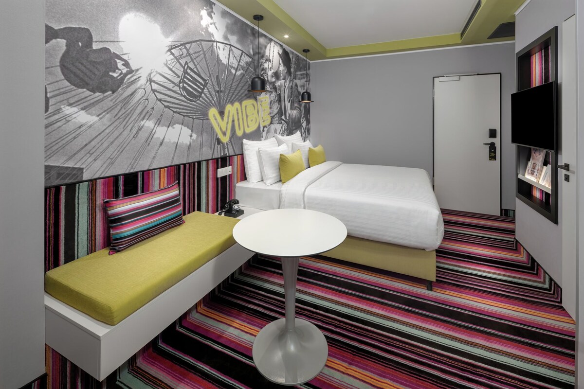 Une chambre d’hôtel avec un lit, une petite table, un banc avec des coussins, une fresque murale et une moquette à rayures.