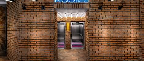 A brick-walled hallway with a neon 'ROOMS' sign, two elevators, and a tiled floor.