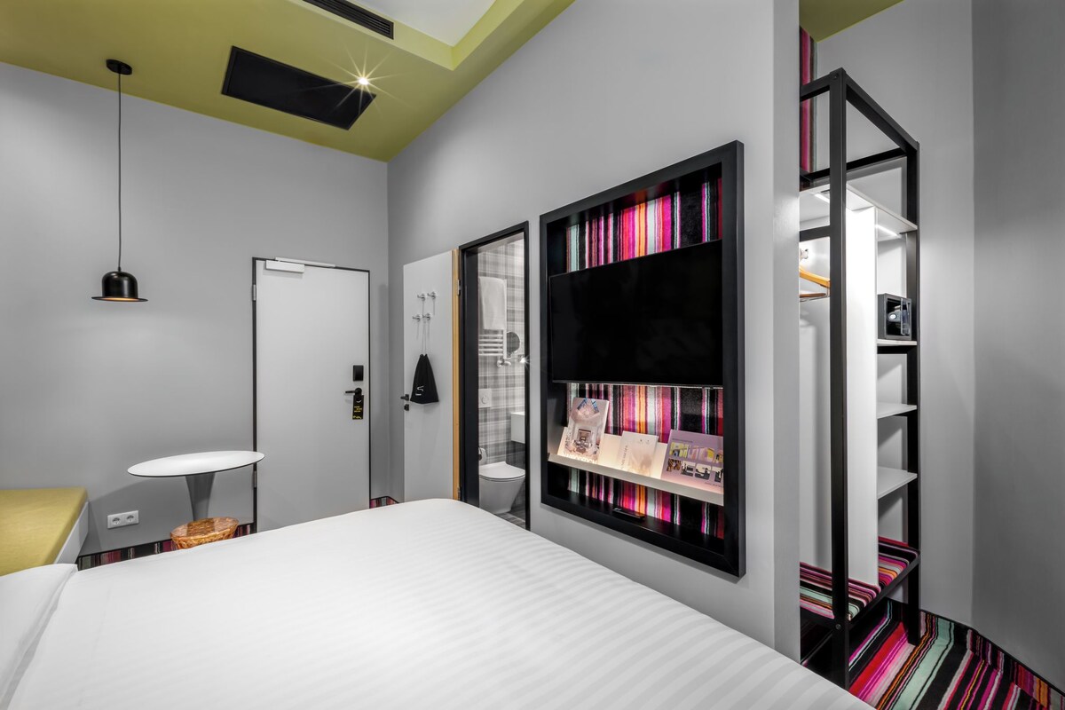 A modern hotel room with a bed, bedside table, lamp, and a built-in bookshelf with colorful items.