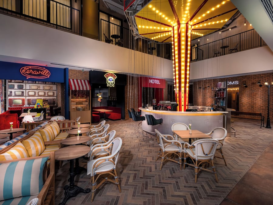 A modern indoor space with a central bar area, seating arrangements, and visible store fronts including a hotel and a store named 'DMS'.