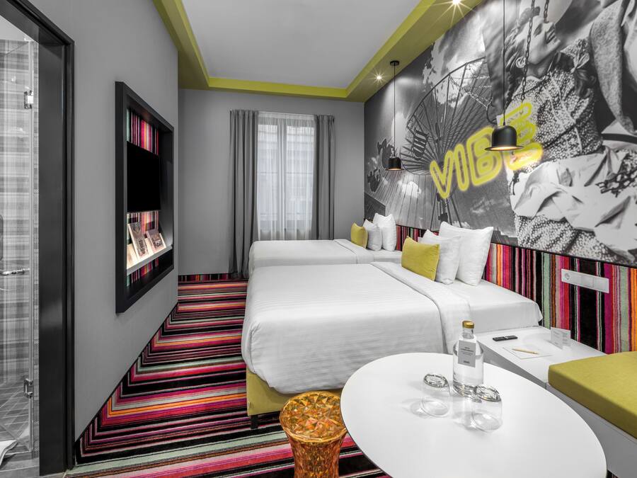 A modern hotel room with a large bed, a small sofa, a round table, and a colorful striped carpet.