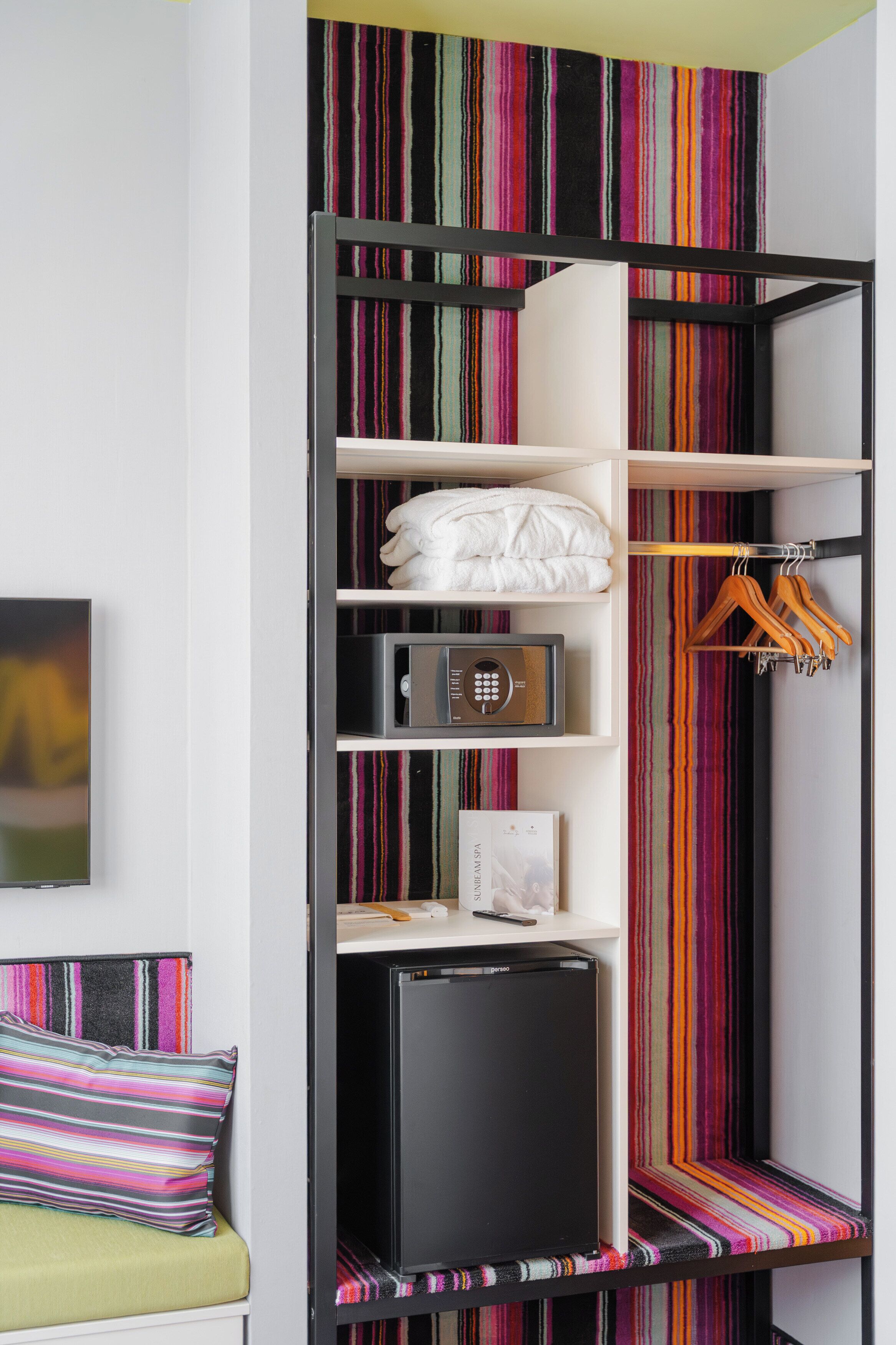 A compact room with a striped wall, a black shelving unit with various items, a small bed with a colorful pillow, and a television on the left.