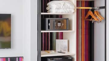 Una habitación pequeña con una pared a rayas, una estantería negra con varios objetos, una cama con un cojín de colores y un televisor a la izquierda.