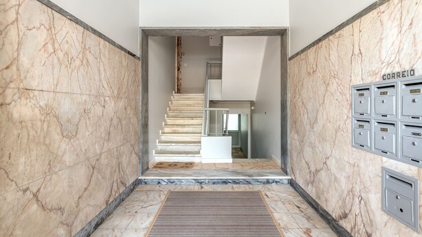 Un pasillo con suelo de mármol y una escalera que lleva a una plataforma, buzones y una rejilla de ventilación en la pared.