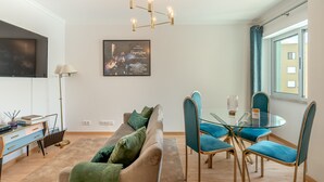 Una sala de estar moderna con un sofá, una mesa de comedor de vidrio y sillas azules. Se observa una araña, un televisor de pantalla plana y un cuadro enmarcado en la pared.