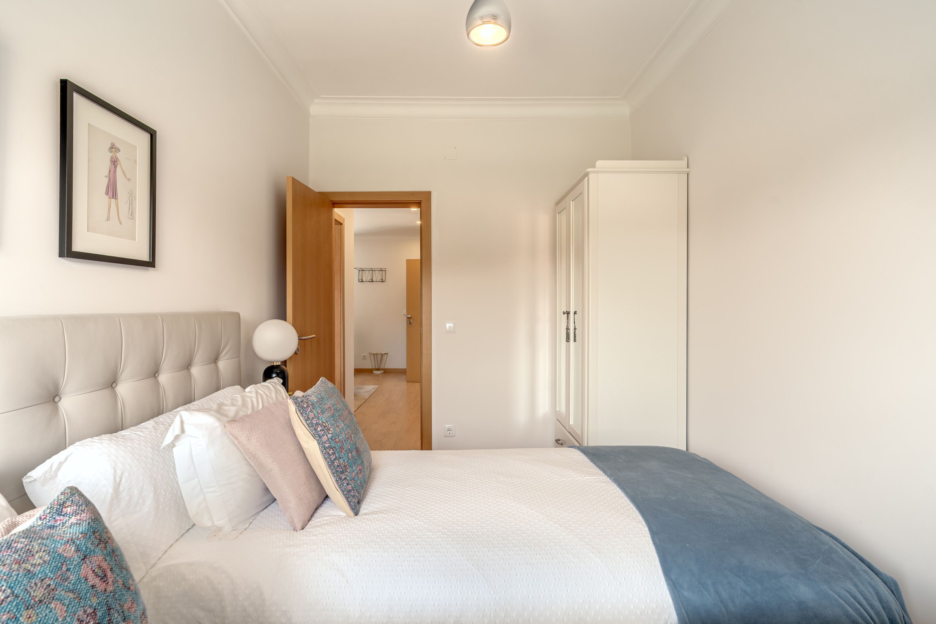 Una camera da letto con testiera capitonata, un letto con biancheria bianca e cuscini a fantasia, un quadro appeso alla parete e un armadio.