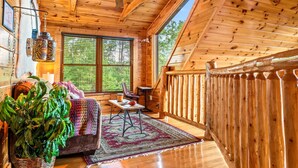 A wooden cabin interior with a ceiling fan, a large window, a wooden deck, a cushioned sofa, a table, and a potted plant. - Mountain Views • Hot Tub • Minutes to Dollywood (Sevierville)