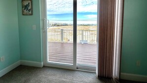 Una habitación con paredes color turquesa, piso alfombrado y puerta corredera de vidrio que da a un balcón con vista a la playa y casas.