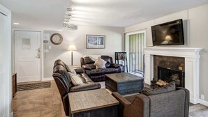 Una sala de estar con un sofá seccional, una mesa de centro, una chimenea, un televisor y una puerta con un aviso.
