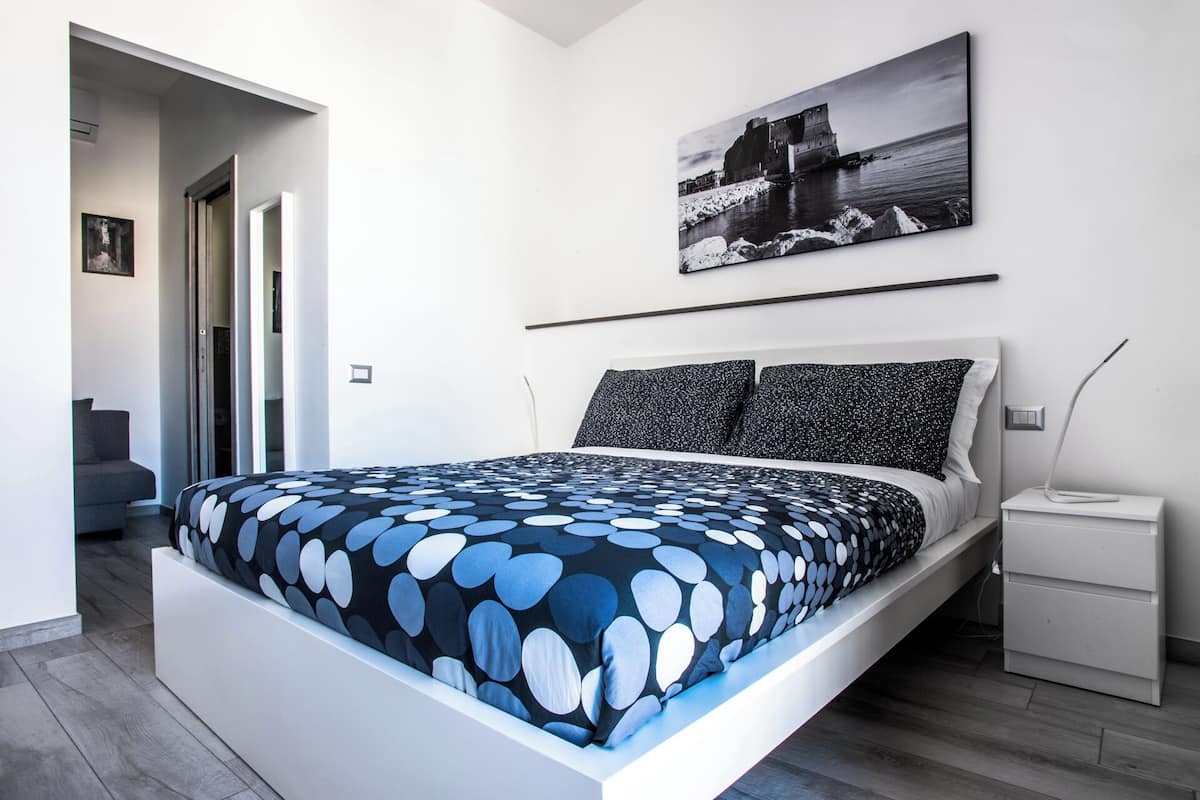 A modern bedroom with a bed, a nightstand, a painting, and a view of the sea.