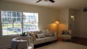 A living room with a sofa, armchair, and floor lamp. There is a window with blinds, a ceiling fan, and a door in the background. - Corner Cottage - 2BR, Close to Lake, Restaurants! (Spring City)