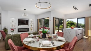 A modern dining area with a round table set for four, a television mounted on the wall, and a view of the outdoors through large sliding doors. - Vila SUNLIFE with pool&sauna by Villas Guide (Matulji)
