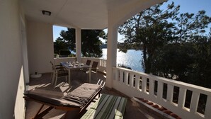 Een balkon met een tafel en stoelen, een comfortabele bank en uitzicht op een meer met bomen.