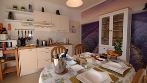 Une cuisine avec une table à manger pour deux personnes, un meuble avec des portes en verre et un mur orné d’une fresque florale aux tons violets.