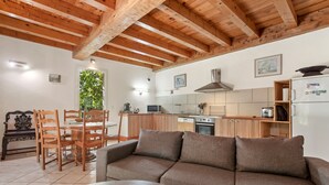 Ein gemütliches Wohnzimmer mit Holzbalkendecke, einer Couch, einem Esstisch mit Stühlen und einer Küchenzeile mit Schränken und Geräten.