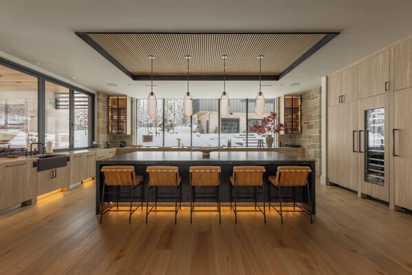 Une cuisine moderne dotée d’un îlot central, de tabourets de bar, de luminaires suspendus et offrant une vue sur un paysage enneigé.
