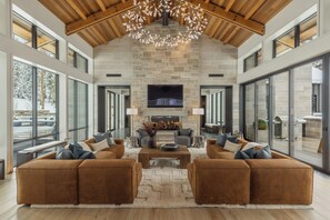 A spacious living room with a stone fireplace, large windows, and a high ceiling with wooden beams. - Brand New Modern Home with Guest House by InvitedHome | Hot Tub, Theater, Gym (Mountain Village)