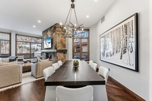 A modern dining area with a long table, white chairs, a fireplace, and a large framed picture on the wall. - Contemporary Condo by InvitedHome | Ski-In/Out, Base of Village, Balcony, Views (Avon)